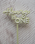 Cake topper with 'Henry is ONE' text on a textured surface